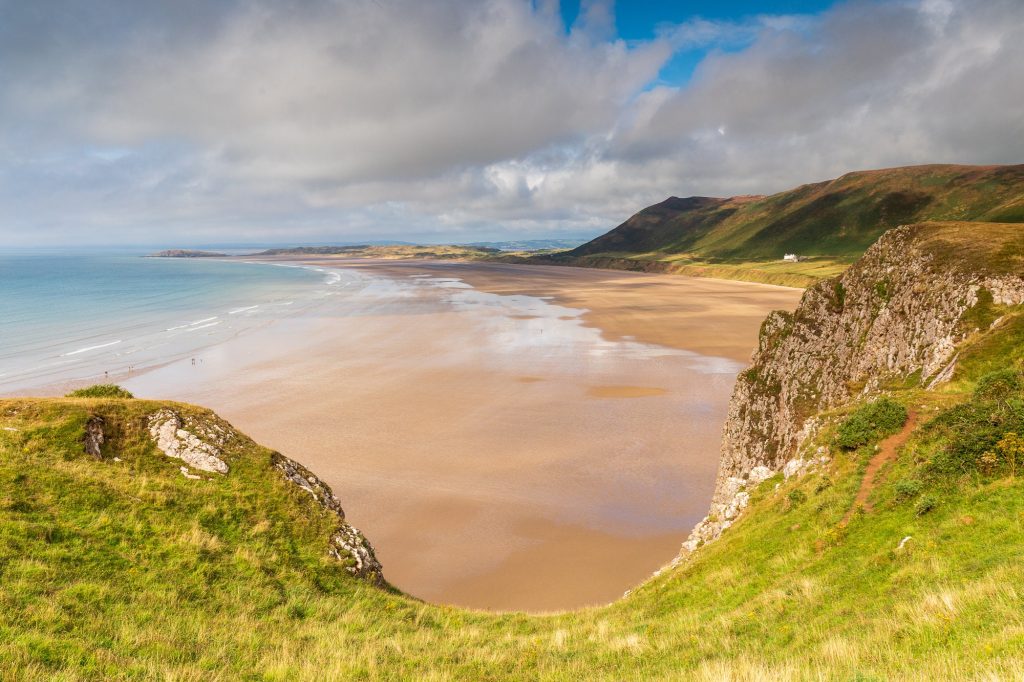 Seen from the cliffs above, Rhossili Beach opens into a wide sweep of golden sand and pale blue sea, Gower – view 2