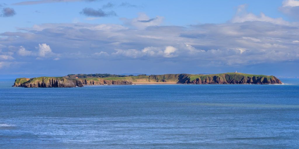 Caldey Island sits low on the horizon beyond the blue waters of the Pembrokeshire coast in this panoramic landscape