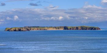 Caldey Island sits low on the horizon beyond the blue waters of the Pembrokeshire coast in this panoramic landscape